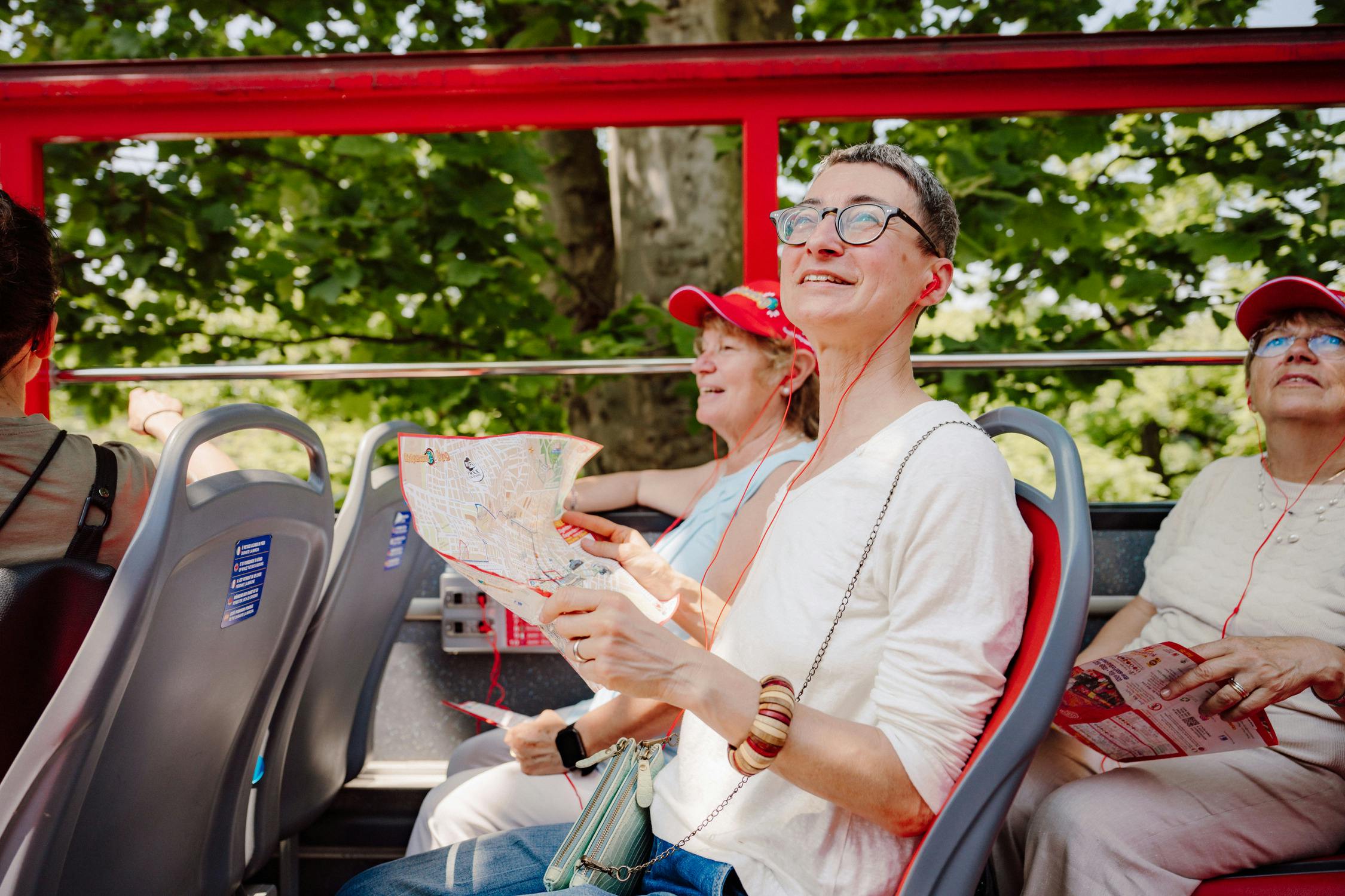 Three people on a double-decker bus tour, wearing headphones and holding maps, surrounded by trees.