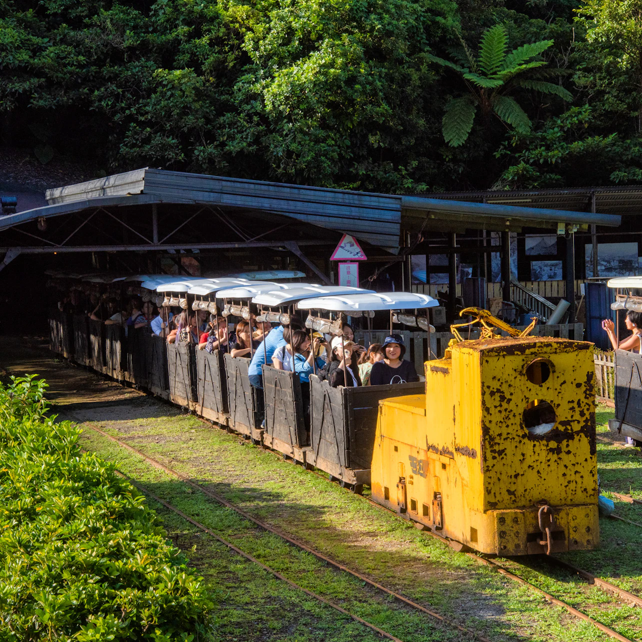 A yellow train with open passenger cars full of people is parked near a station surrounded by lush greenery.