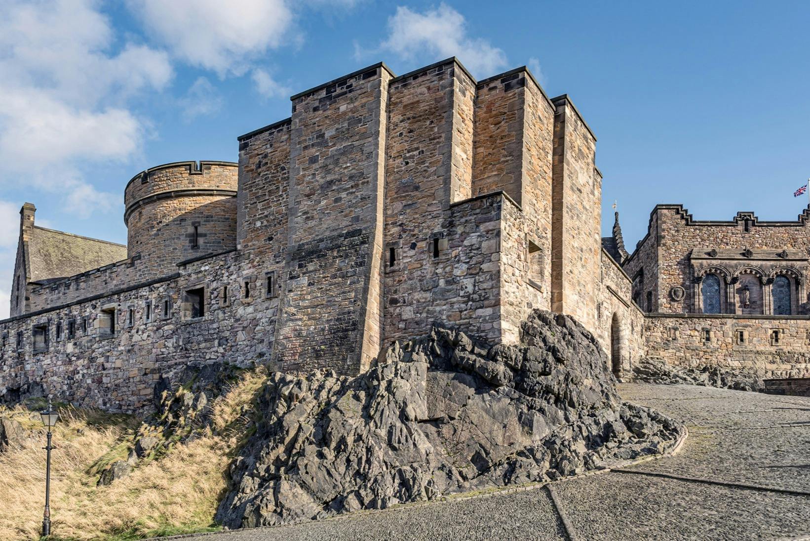 Fortezza di pietra con alte mura e torrette su una collina rocciosa sotto un cielo blu con nuvole.
