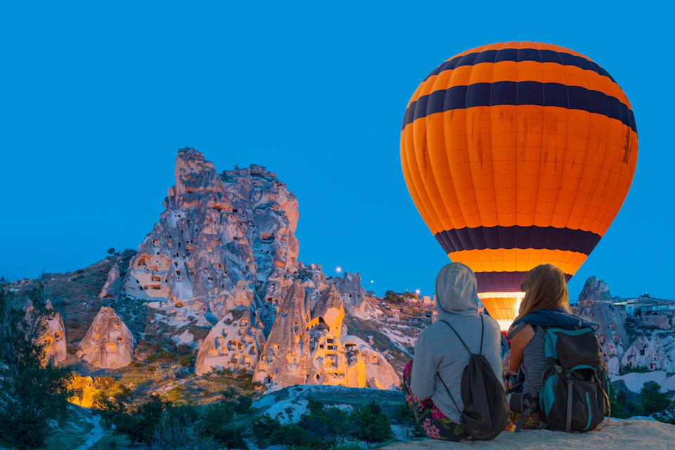Two people sit on a ledge at dusk, watching a hot air balloon near rock formations with cave dwellings.