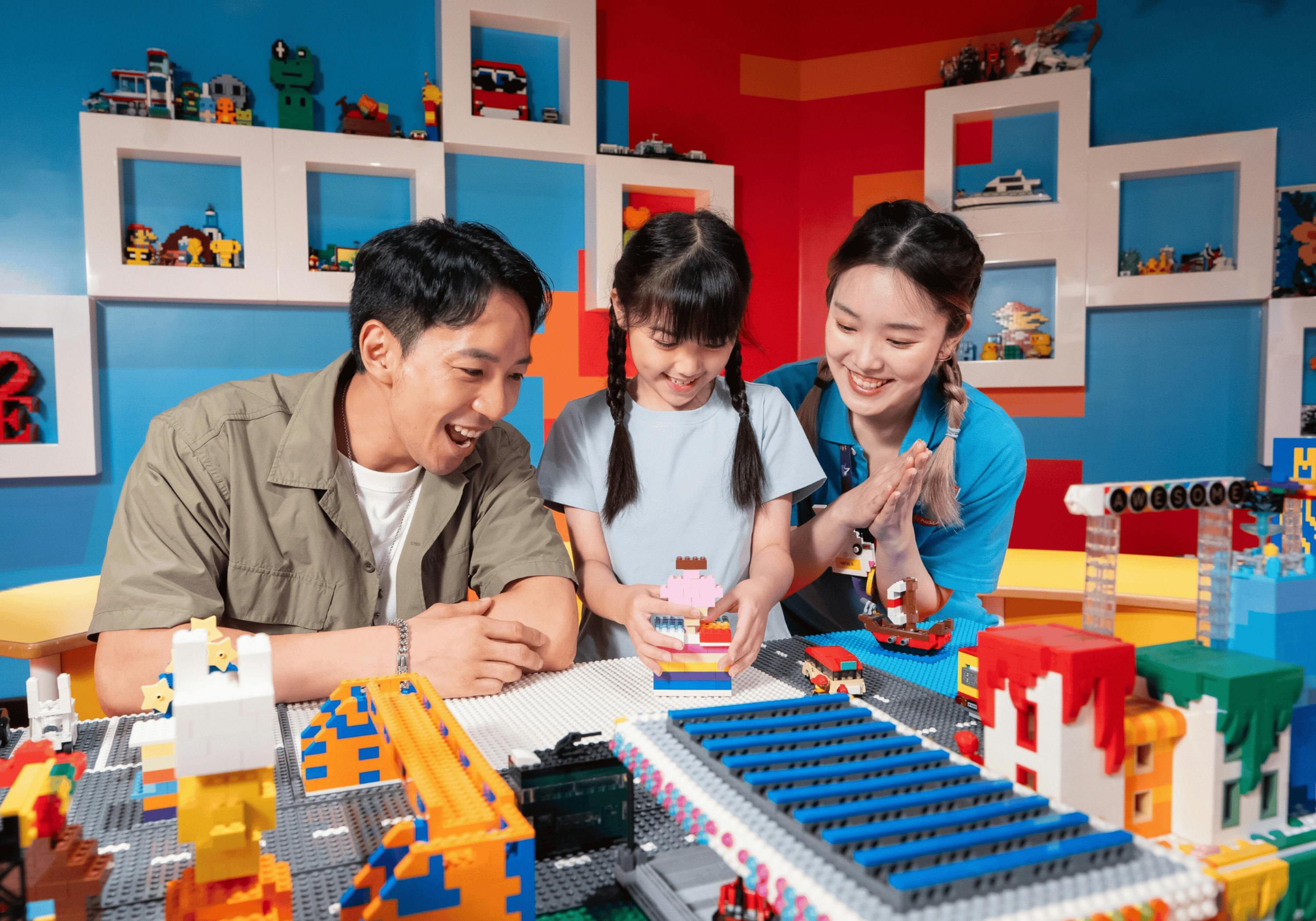 A child and two adults building with colorful bricks in a vibrant playroom with shelves displaying various models.