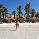 Una persona si fa strada nell'acqua bassa della spiaggia verso capanne e palme sotto un cielo azzurro e limpido.