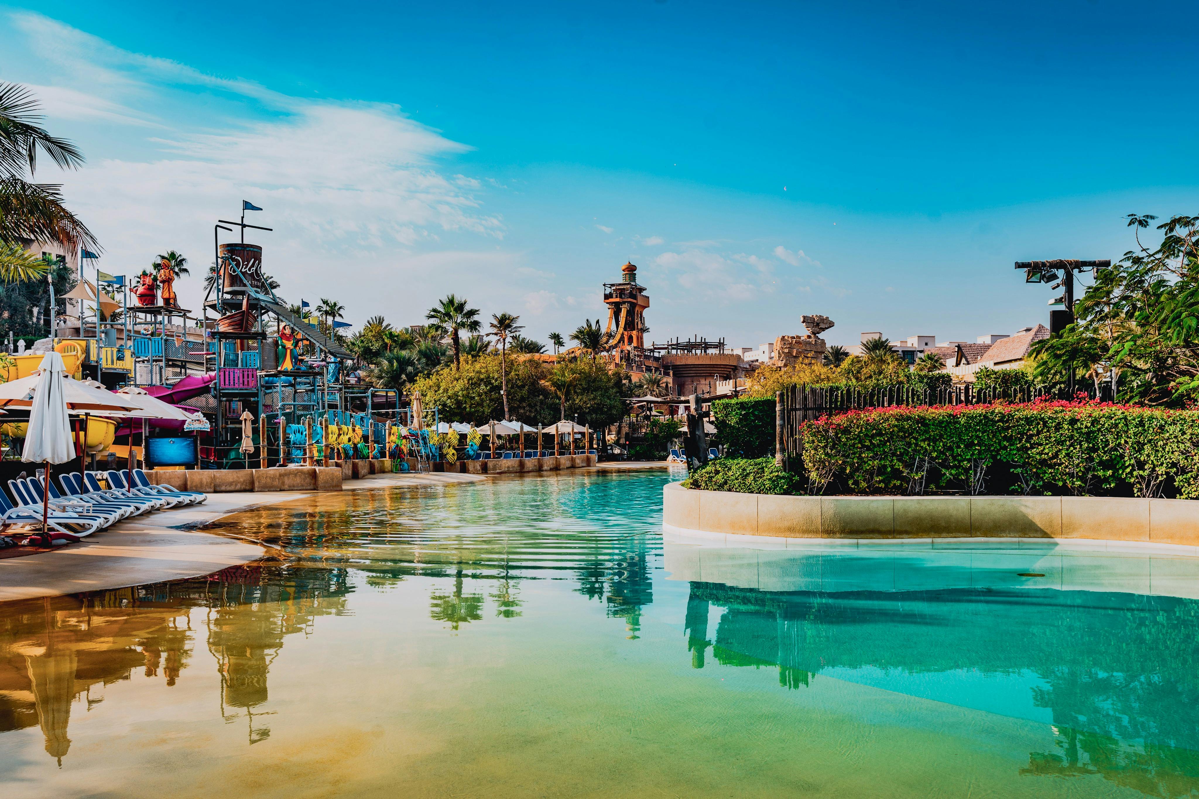 Un parc aquatique avec des toboggans, une rivière tranquille, des plantes tropicales et des structures sous un ciel bleu clair.