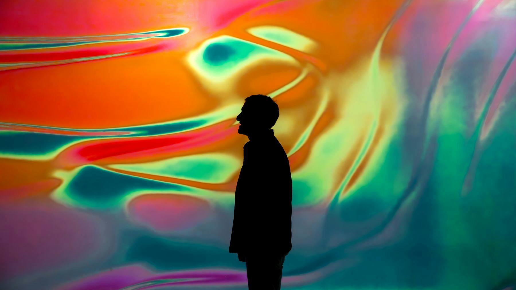 소용돌이치는 주황색, 청록색, 분홍색의 다채롭고 추상적인 배경에 서 있는 사람의 실루엣.