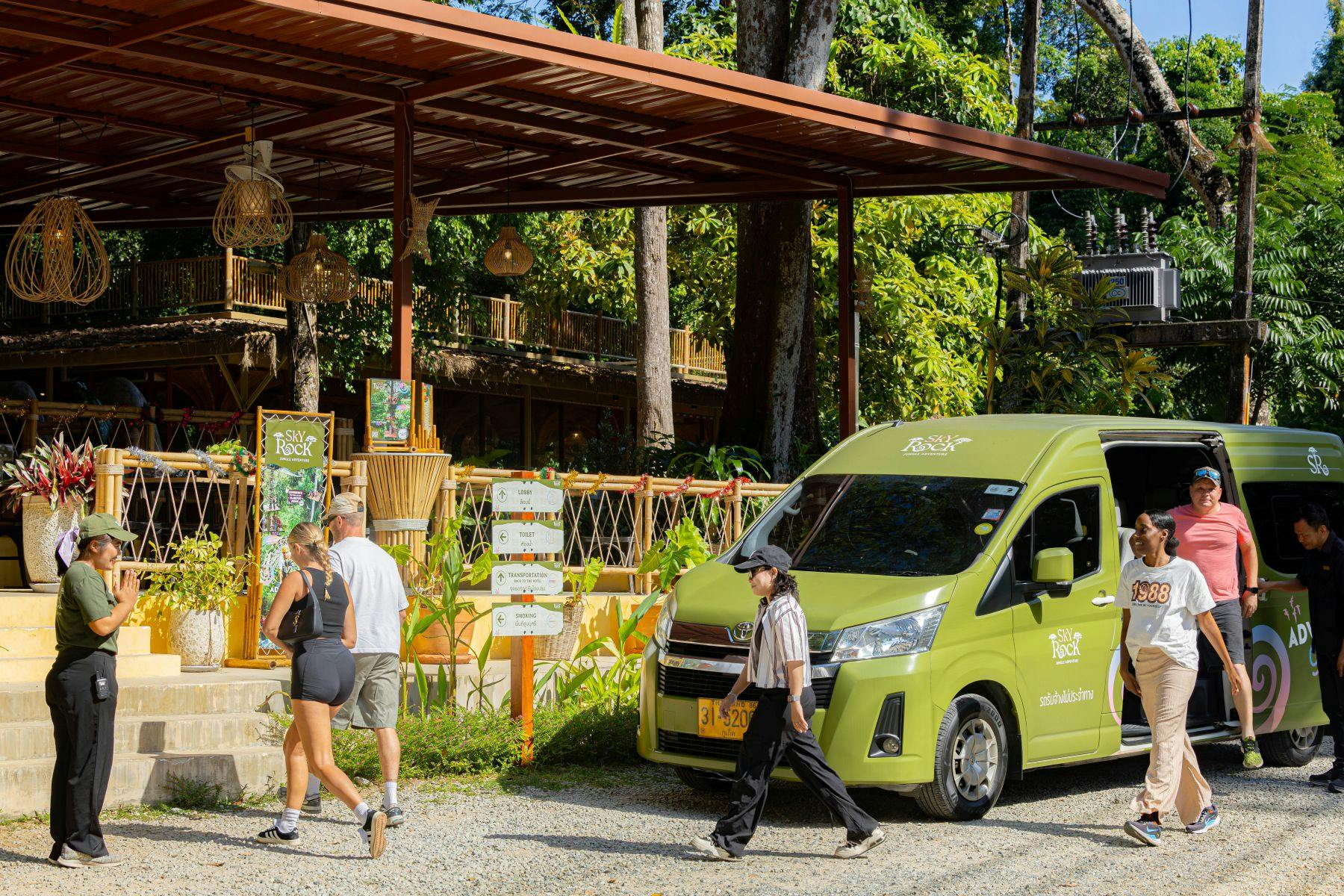 A green van labeled 'The Rock' parked near a tropical outdoor setting with people walking by and trees in the background.