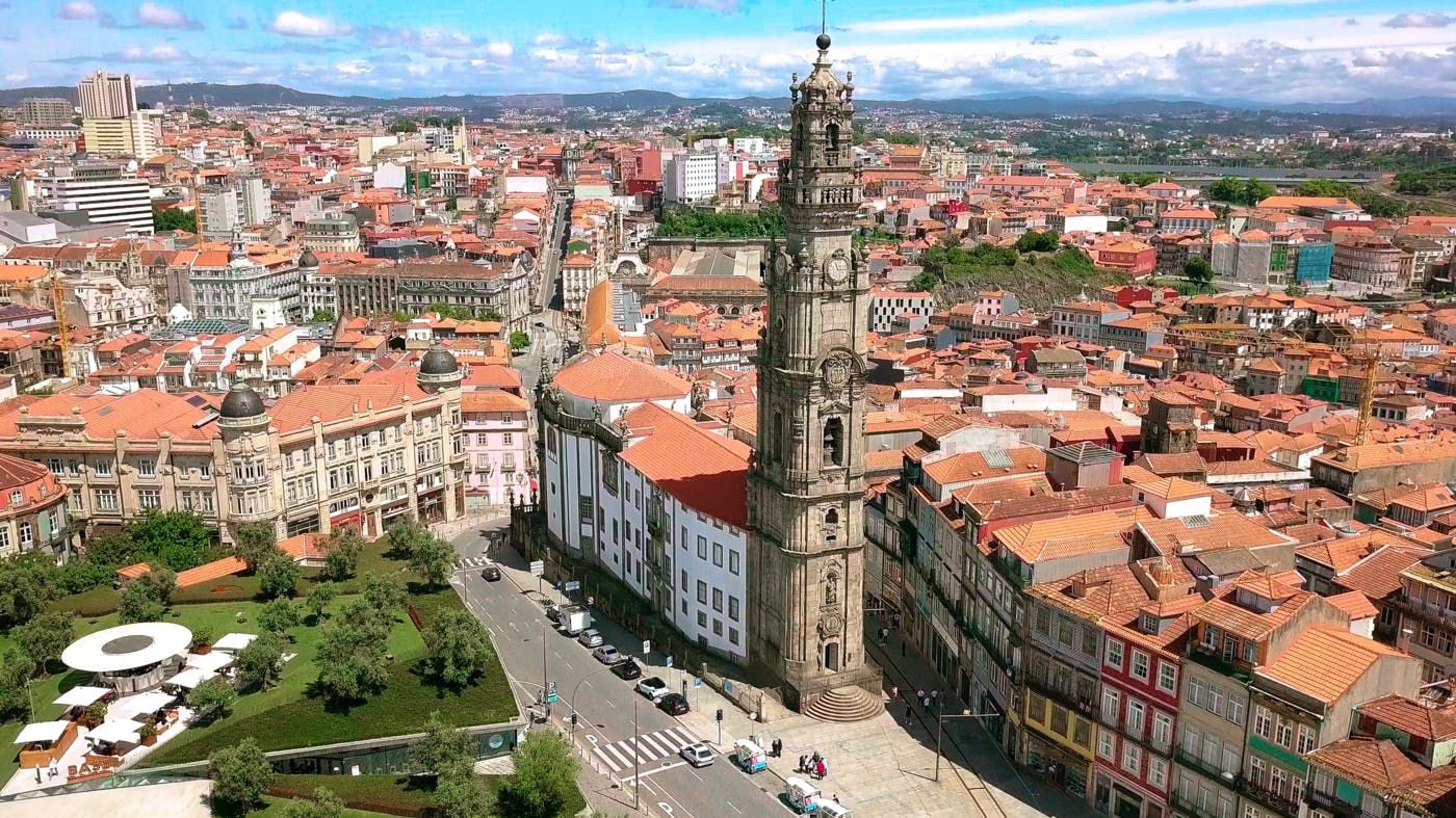 Letecký pohled na panorama města s výraznou hodinovou věží, historickými budovami, střechami z červených tašek a zeleným parkem.