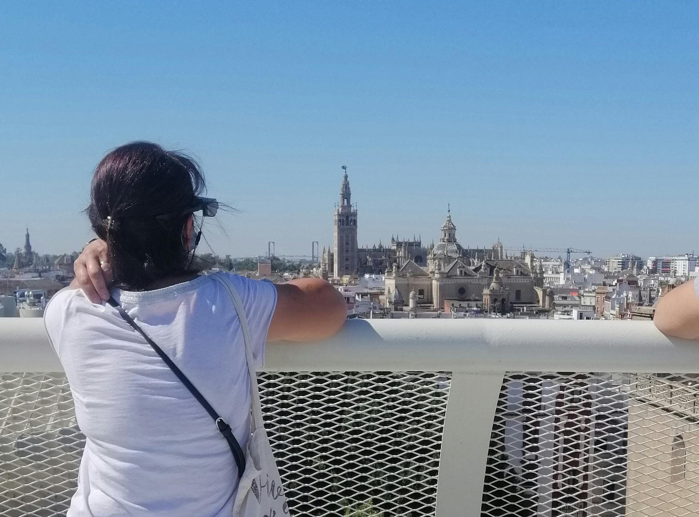 Ένα άτομο με σκούρα μαλλιά και γυαλιά ηλίου στέκεται δίπλα σε έναν φράχτη, κοιτάζοντας ένα αστικό τοπίο που κυριαρχείται από ιστορικά κτίρια και πύργους.