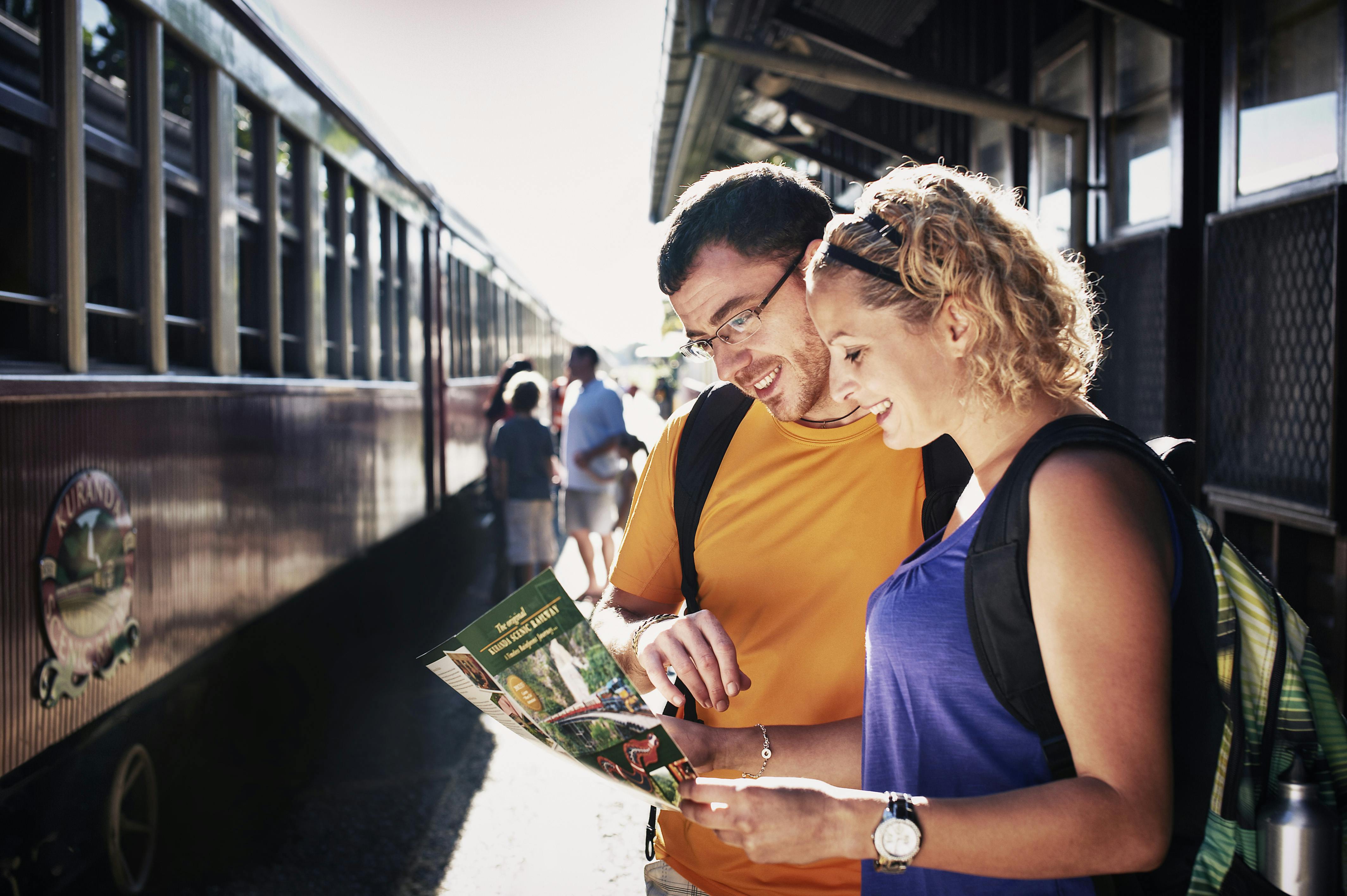 Two people with backpacks, smiling, look at a pamphlet on a train platform with a train and other passengers in the background.