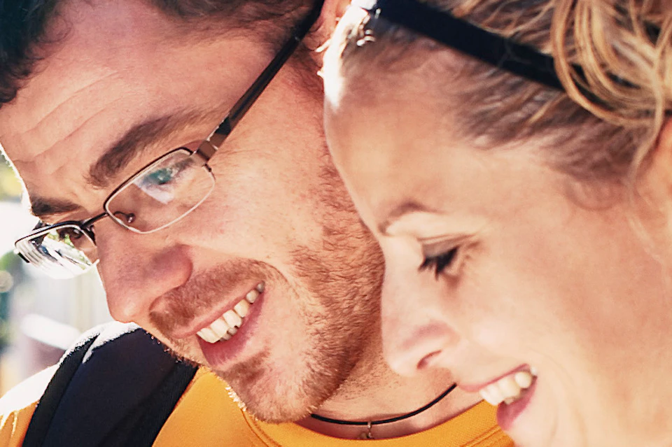 Two people with backpacks, smiling, look at a pamphlet on a train platform with a train and other passengers in the background.
