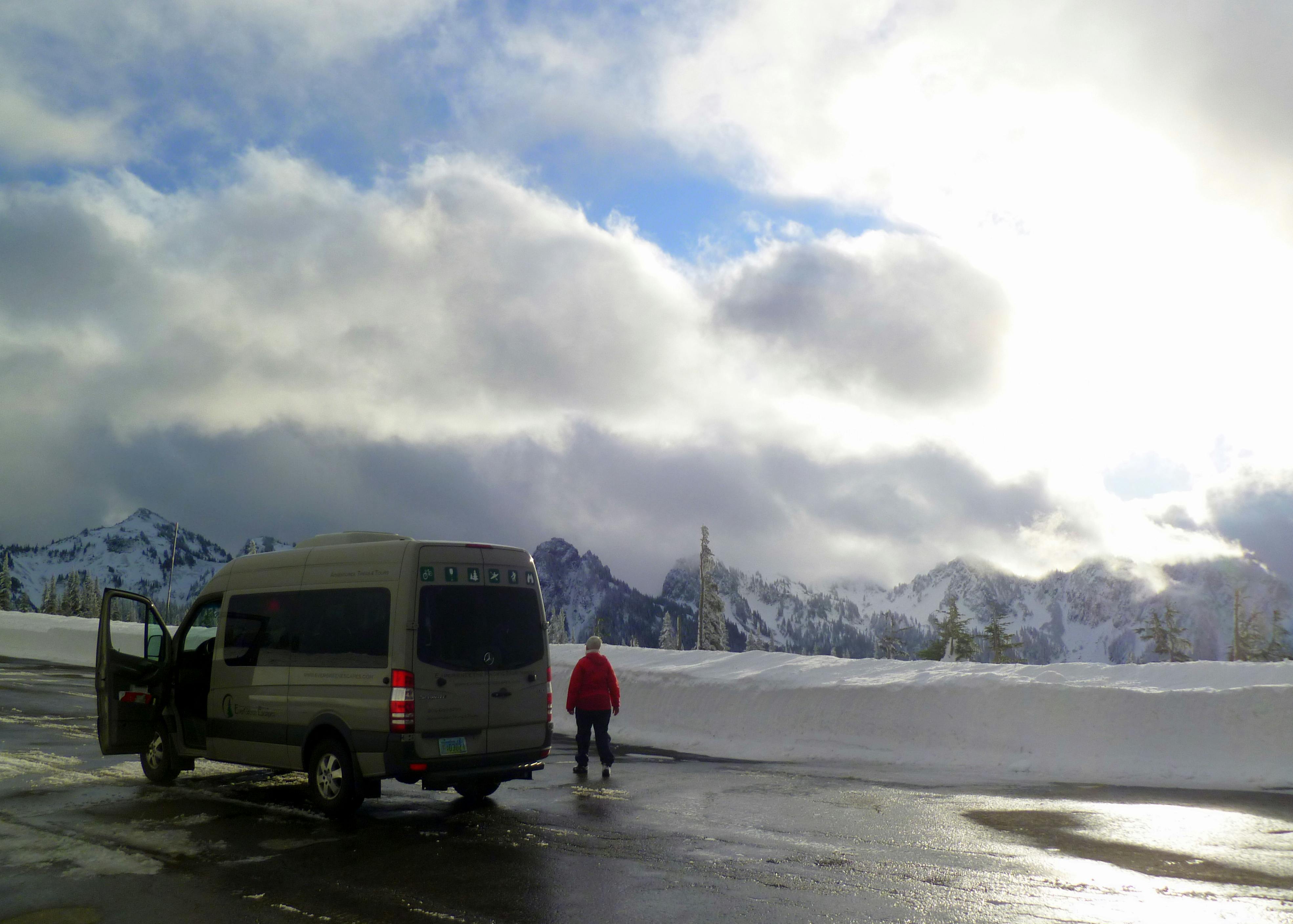 一个穿着红夹克的人站在湿路上一辆停着的面包车旁边，背景是白雪覆盖的山脉和多云的天空。
