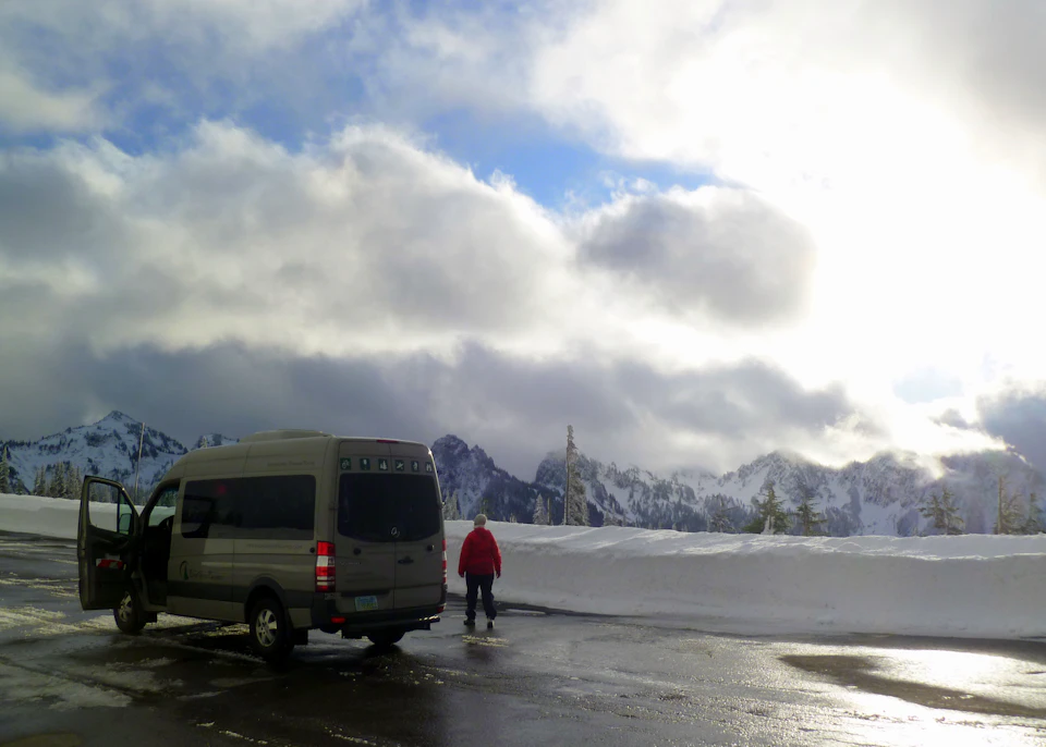 A person in a red jacket stands near a parked van on a wet road, with snow-covered mountains and a cloudy sky in the background.