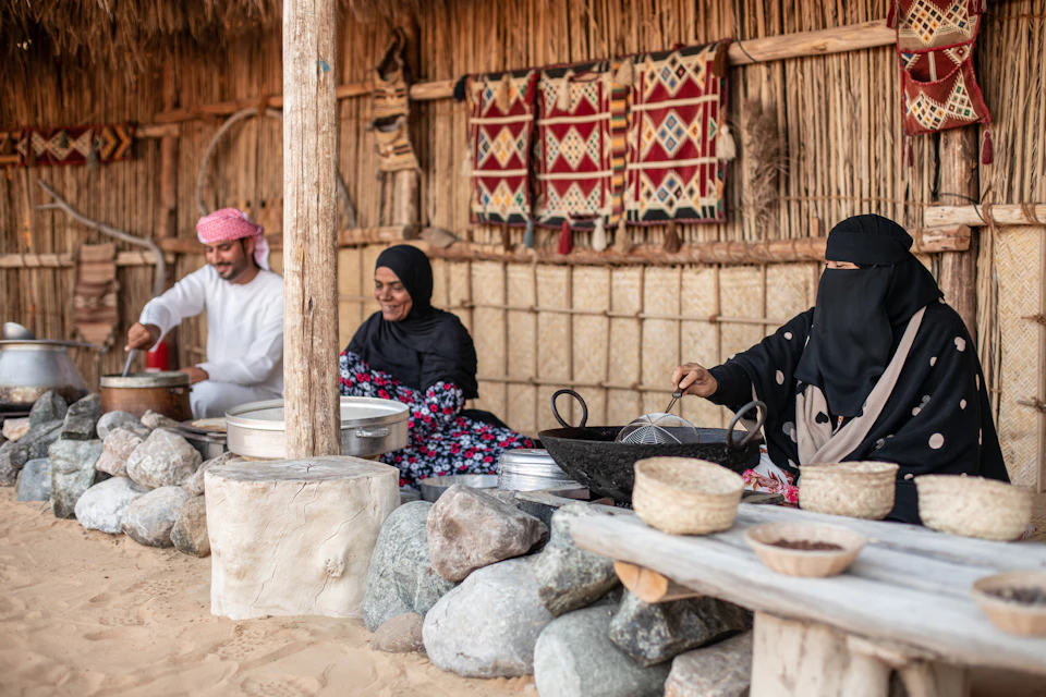 A man and a woman in traditional attire prepare food in an outdoor kitchen with stone and woven mat walls adorned with textiles.