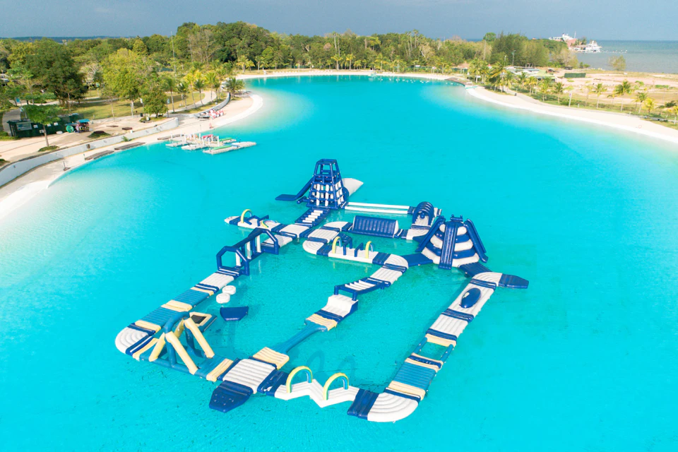 An inflatable water obstacle course floating on a large turquoise lagoon surrounded by trees and a sandy beach.