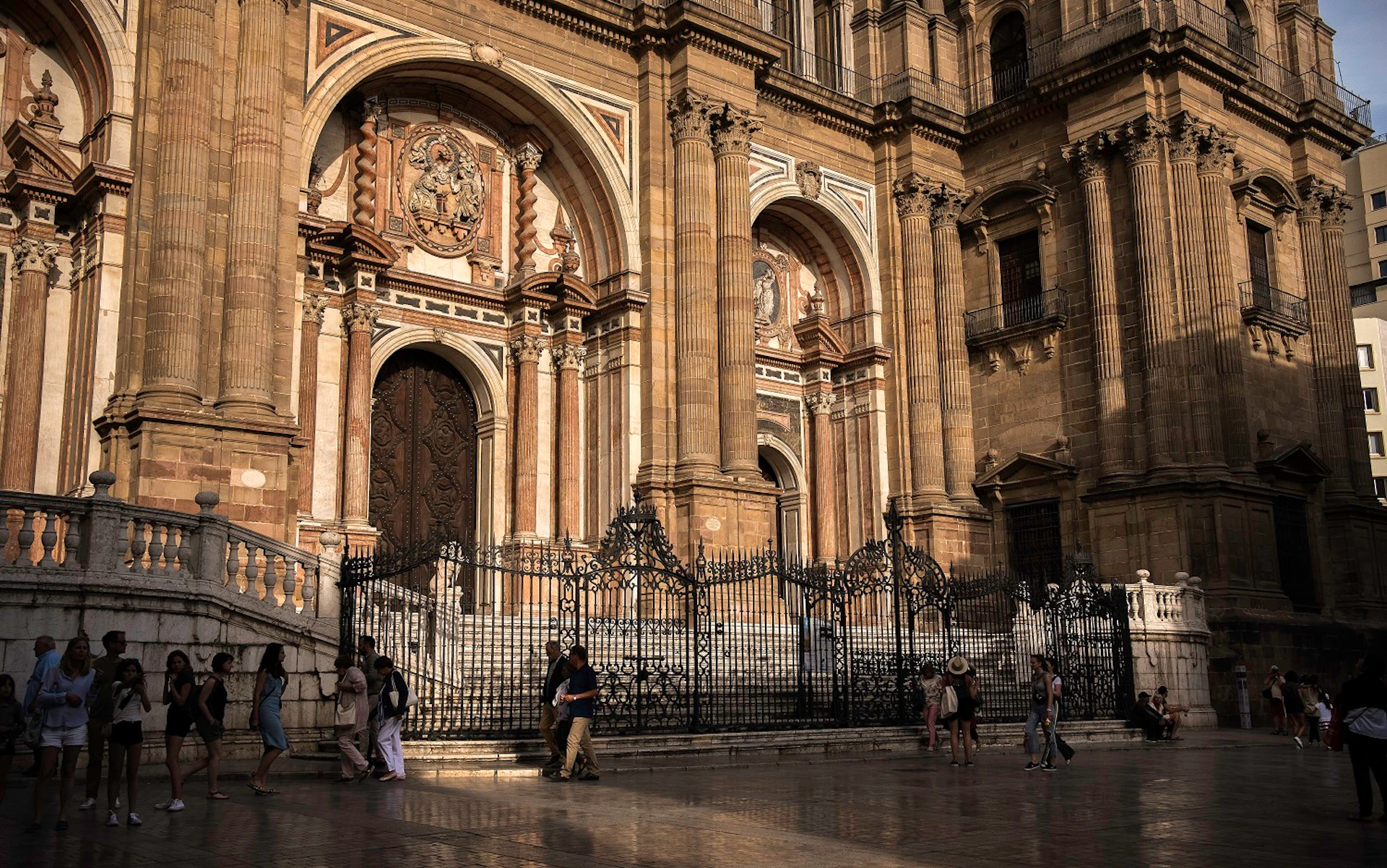People walking in front of a large, ornate cathedral entrance with columns, decorative arches, and an iron fence.
