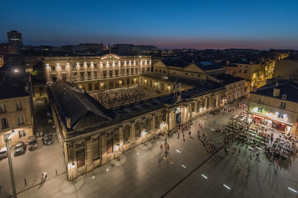 Aerial view of a large courtyard surrounded by historic buildings, with people gathered, illuminated by streetlights at dusk.