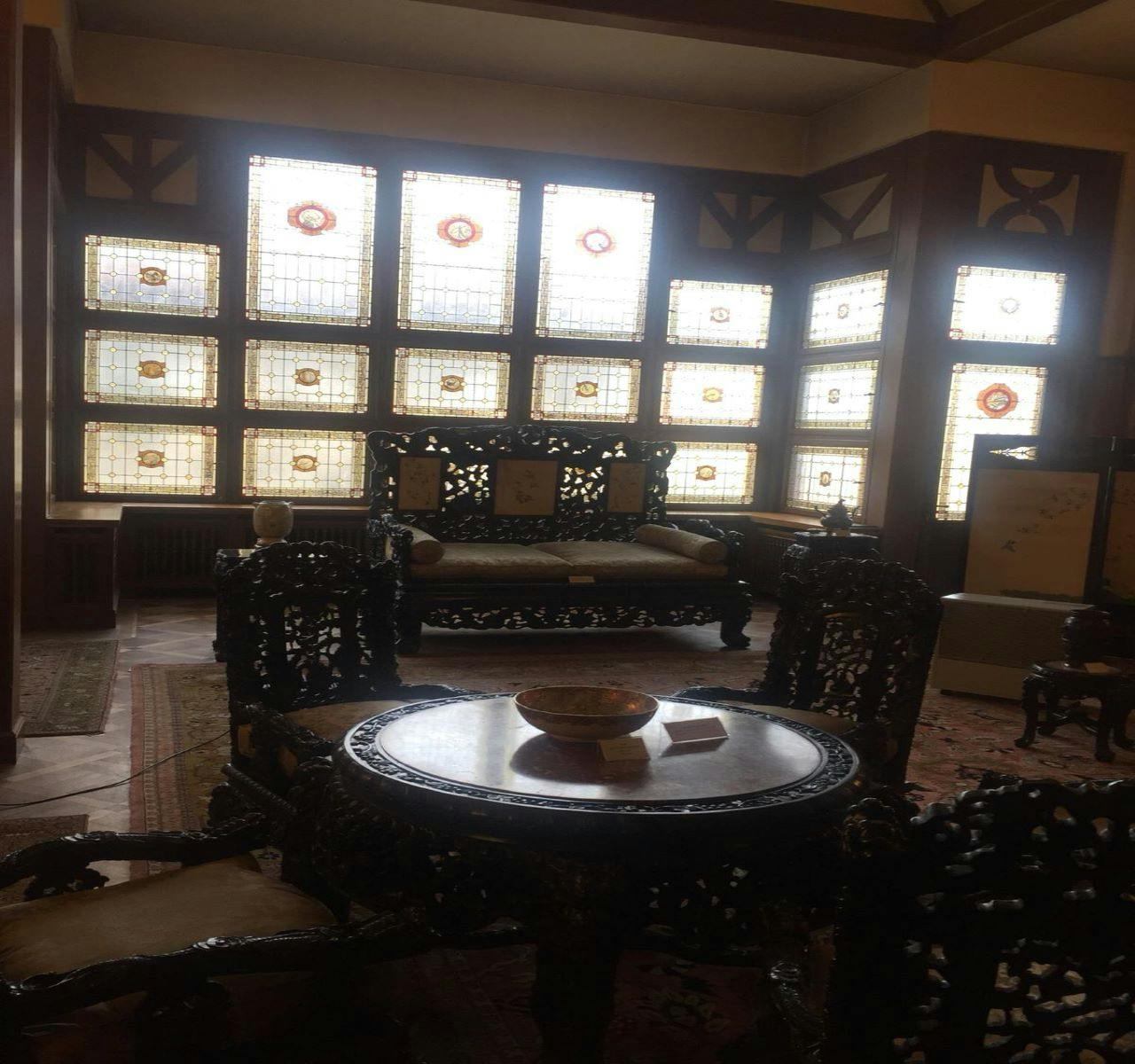 アンティークの木製家具は、複雑なデザインの装飾的なステンドグラスの窓がある薄暗い部屋にあります。