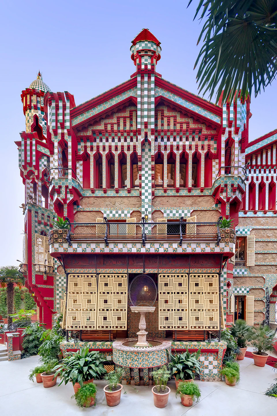 A multicolored, intricately designed building with geometric patterns, a balcony, a fountain, and surrounding greenery.