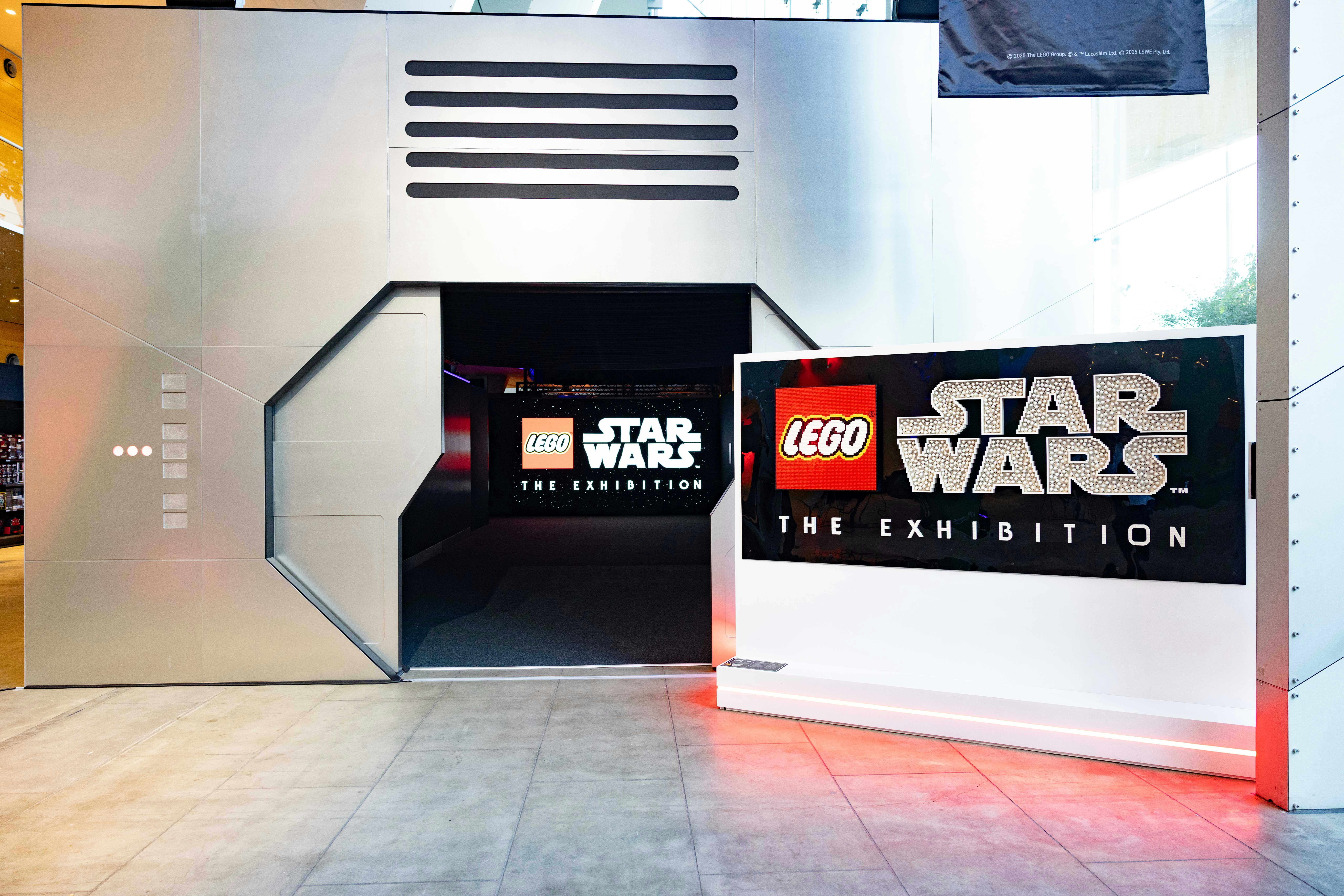 Entrada a una exposició de Lego Star Wars amb un disseny metàl·lic futurista i cartells il·luminats promocionant l'esdeveniment.