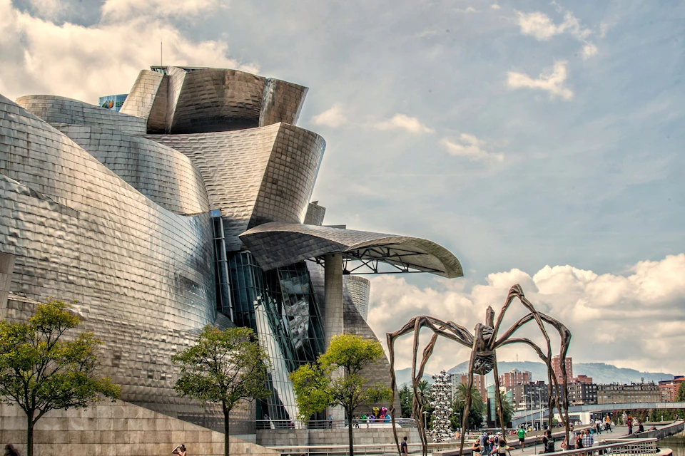 Modern, metallic building with curved architecture. Large spider sculpture and people walking nearby. Trees and a cloudy sky in the background.