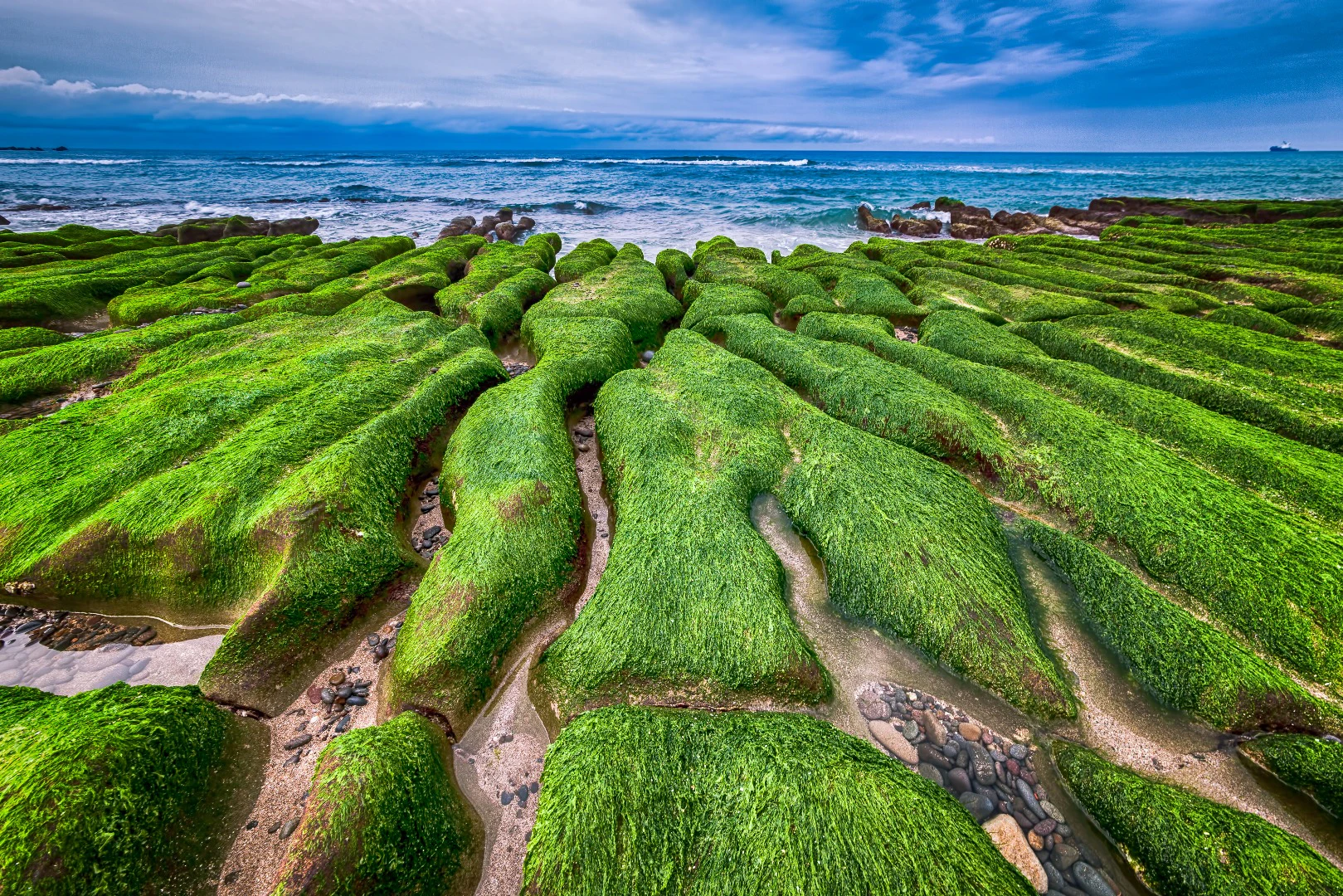 A rocky shore covered in lush green moss, with the ocean and waves visible under a cloudy blue sky.
