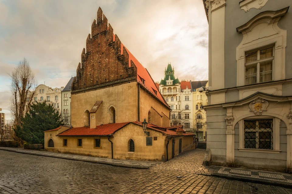 Historical building with a red roof and Gothic elements on a cobblestone street, surrounded by ornate, colorful buildings.