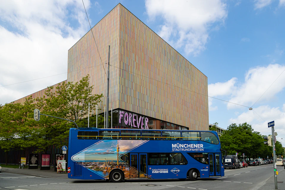 A blue double-decker tour bus in front of a modern building with "FOREVER" displayed on its facade. Trees and a cloudy sky are in the background.