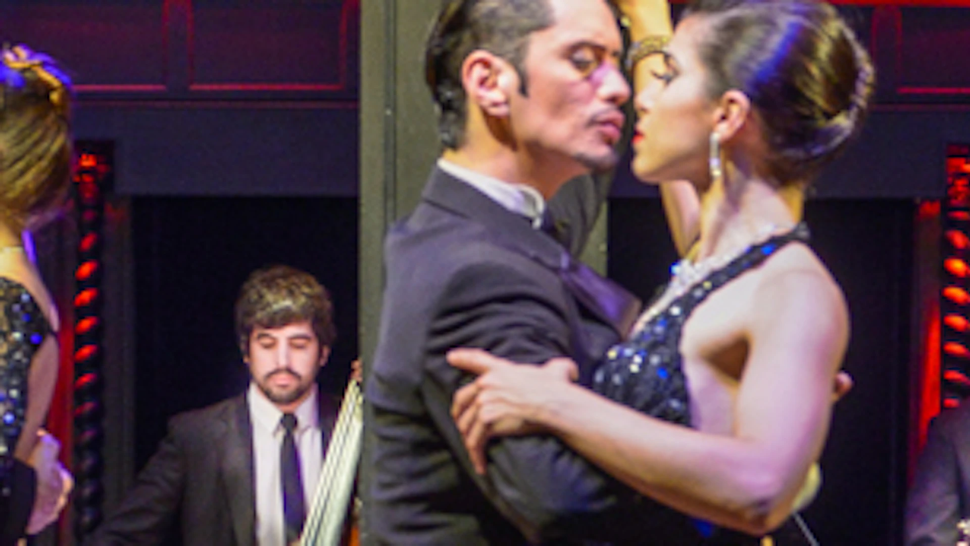 Couples perform a tango on stage in elegant attire, with a band playing in the background. The scene is lit with vibrant colors.