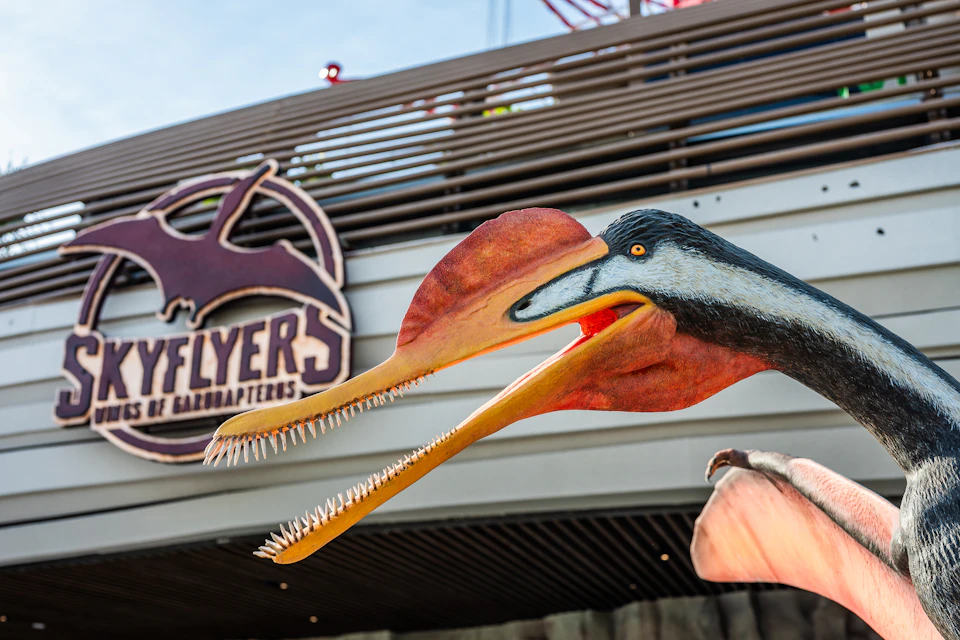 Close-up of a pterosaur sculpture with an open mouth, near a sign reading "Flyers." Background includes a building facade.