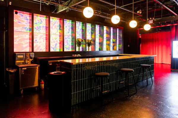 Moderne bar med flisebelagt bord, barstole og oplyste flerfarvede abstrakte paneler på væggen. Runde lamper hænger fra loftet.