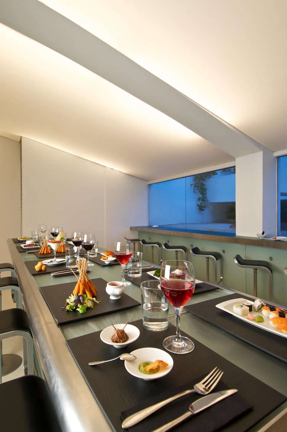 A modern dining table set with various gourmet dishes, wine glasses, and empty seats, under soft lighting near a window.
