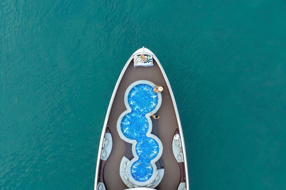 Aerial view of a yacht's bow with a unique, multi-section swimming pool and two people lounging by it in clear blue waters.