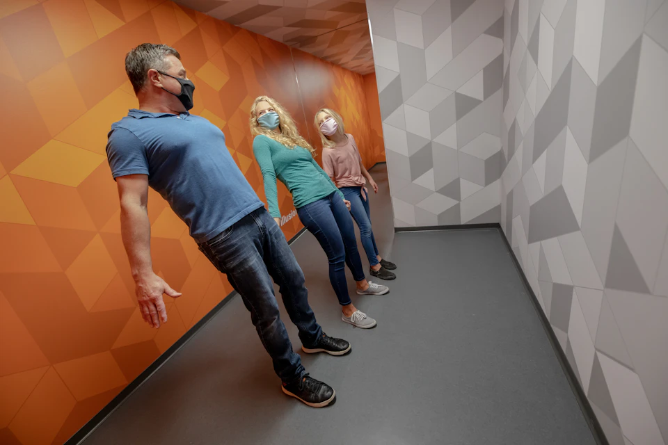 Three people in masks lean against an orange and grey geometric wall, creating an optical illusion in a tilted room.