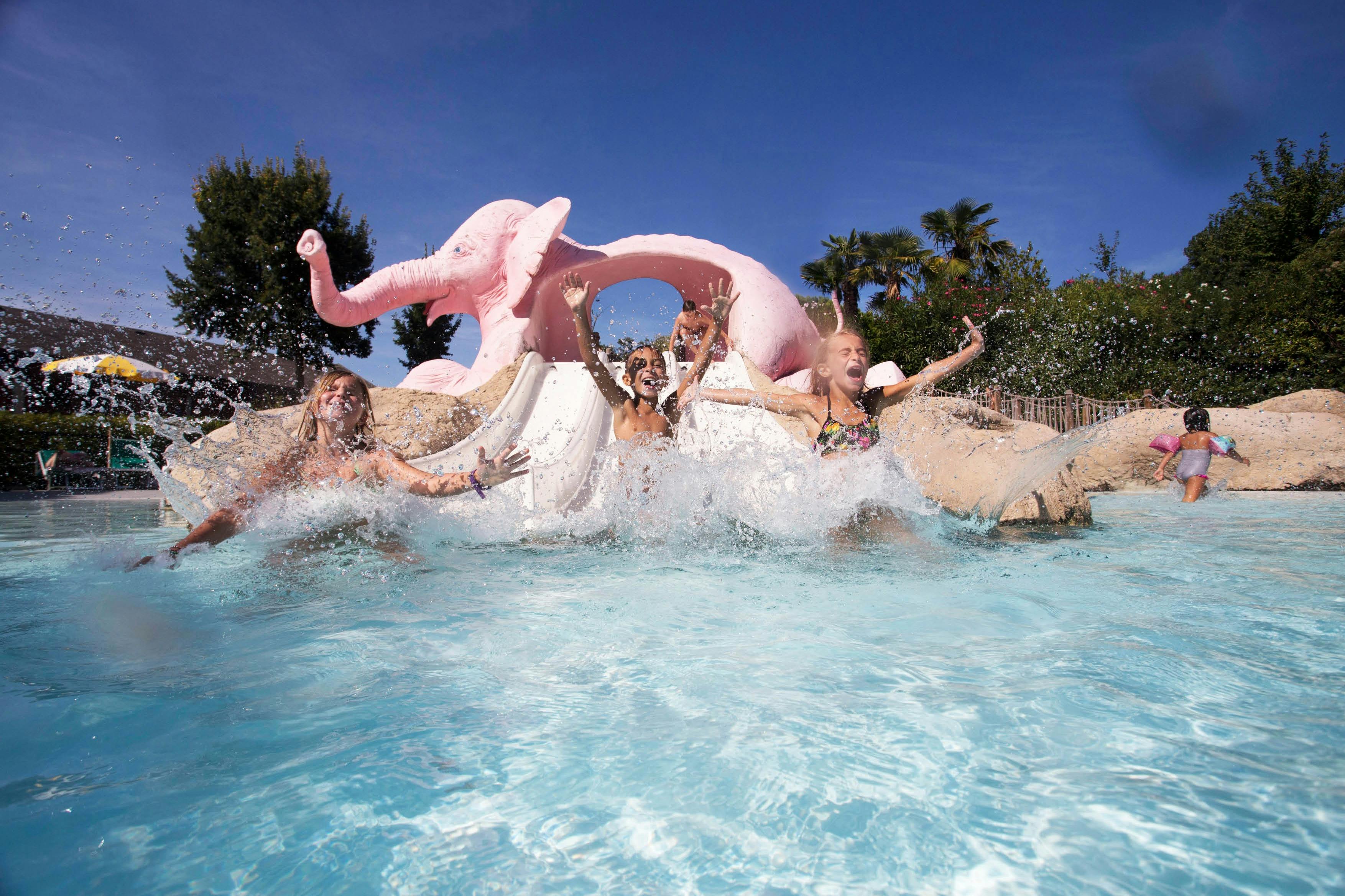 Bambini che scivolano in una piscina da uno scivolo a forma di elefante rosa, spruzzando acqua sotto un cielo azzurro e limpido.
