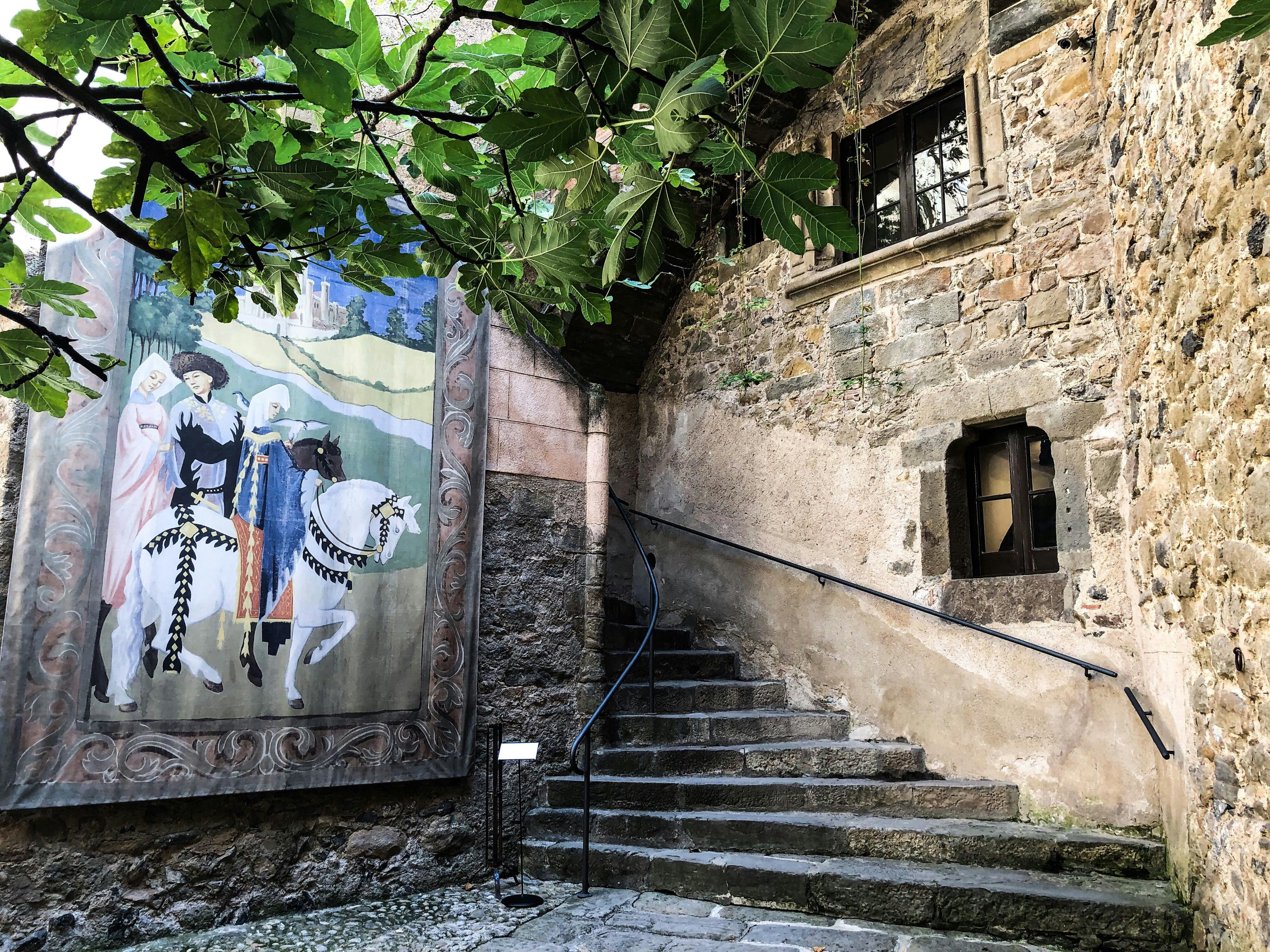 馬に乗った2人の人物の壁画の隣にある石造りの階段。上にはイチジクの木の枝があり、石の壁には窓があります。