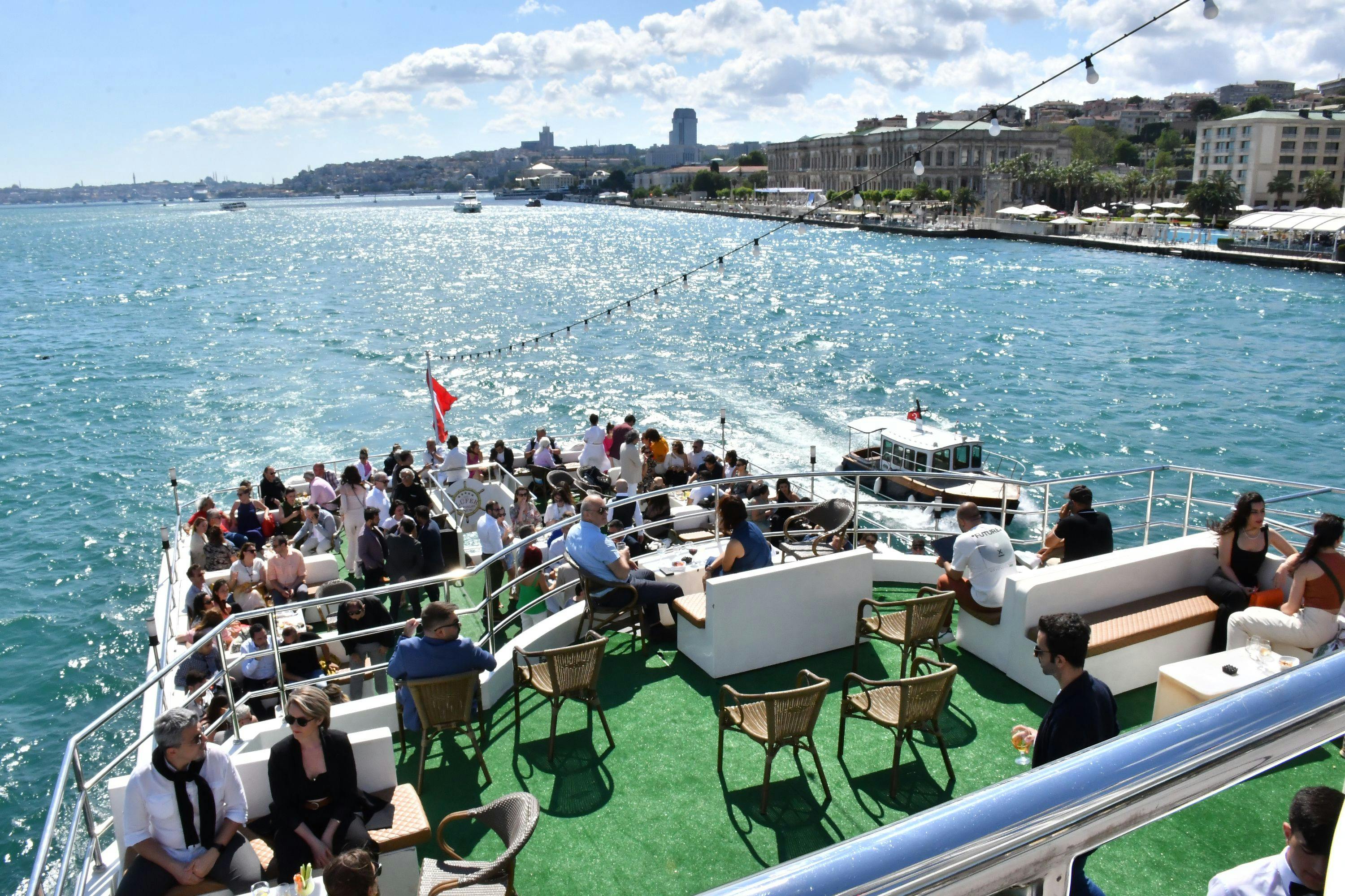 People enjoying a sunny day on the deck of a boat with seating, sailing on blue water near a city shoreline.