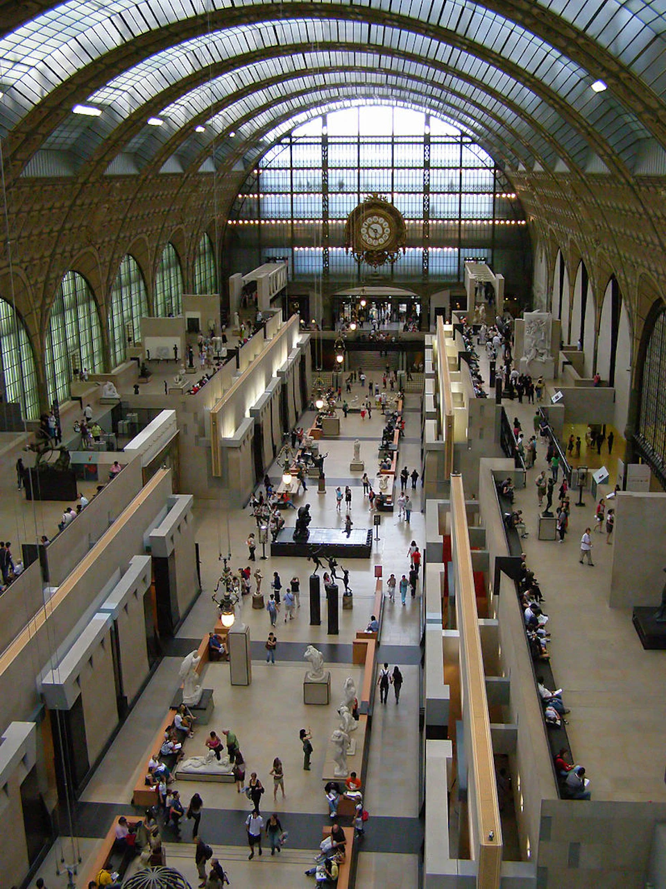 A spacious museum with high arched ceilings, artworks, sculptures, and people walking and viewing exhibits.