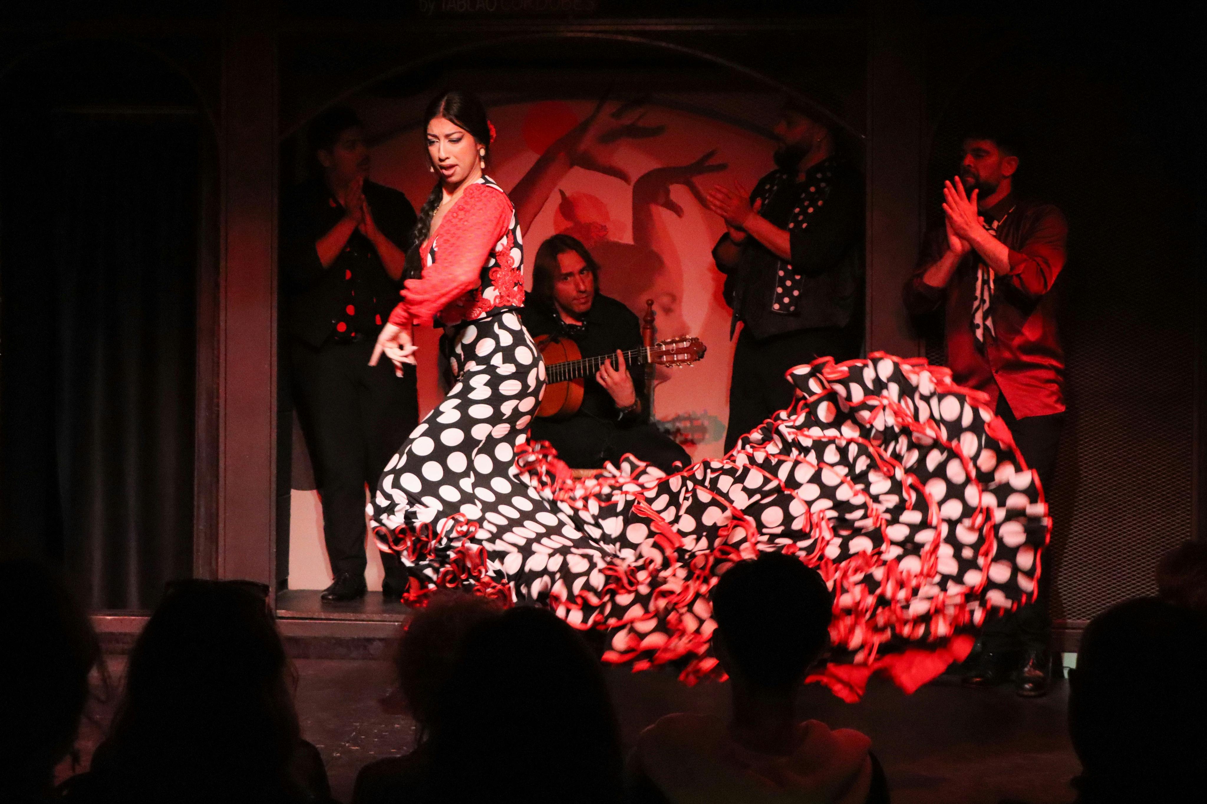 Een flamencodanseres in een rode en zwarte stippenjurk treedt op op een podium met muzikanten die gitaar spelen op de achtergrond.