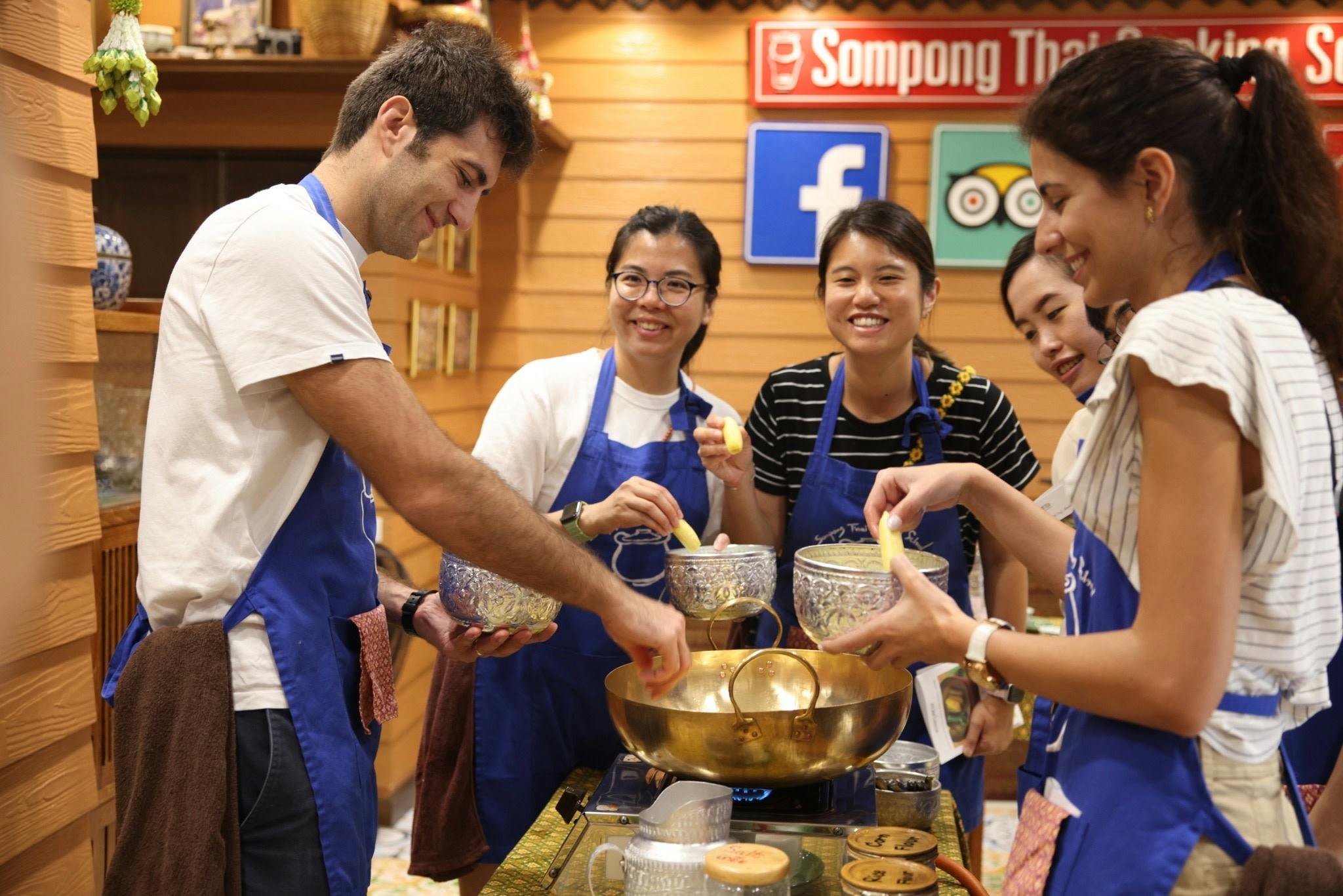A group of people wearing blue aprons participate in a cooking class, preparing ingredients around a golden pot.