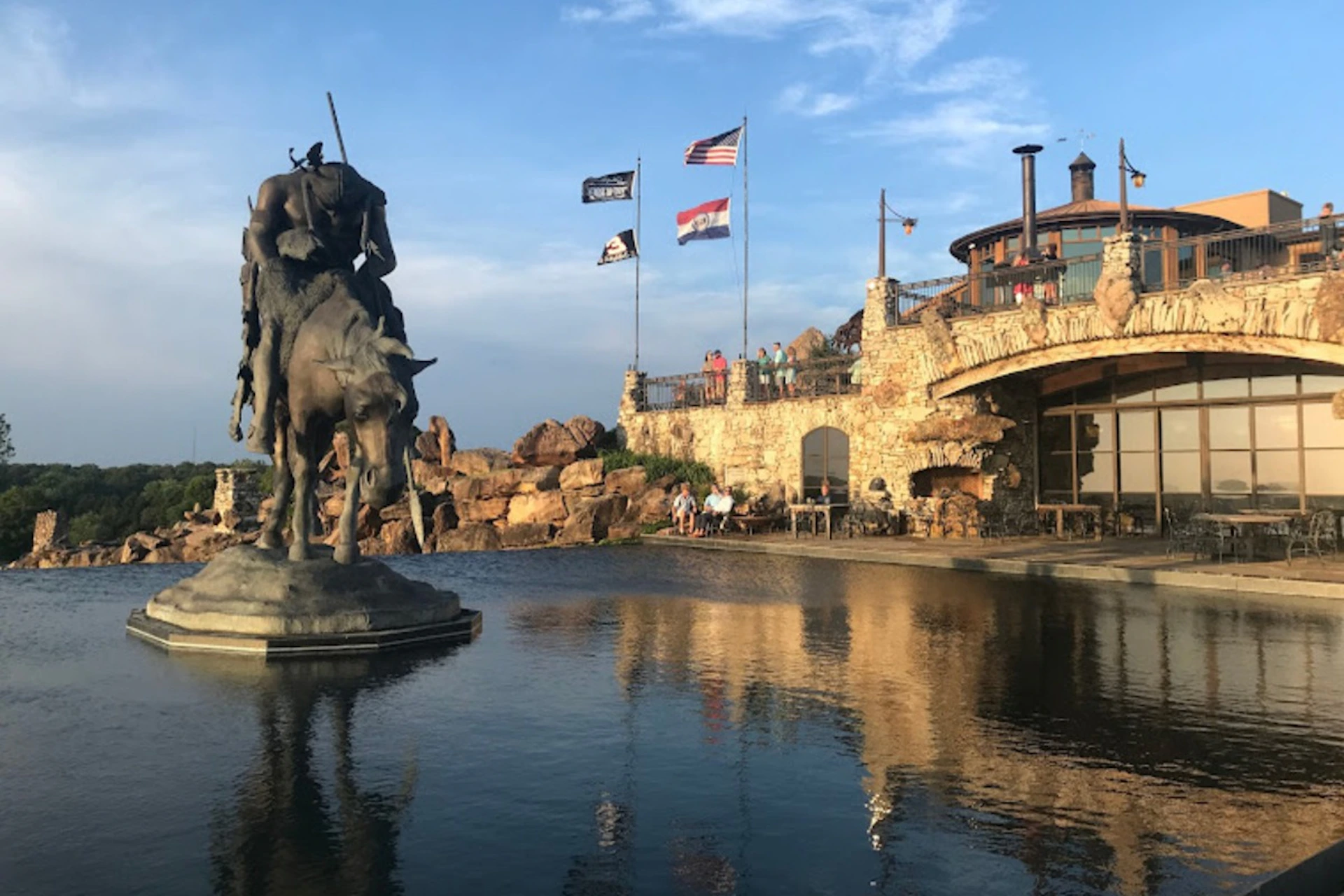 A statue of a rider on a horse stands in a reflective pool. Flags and a stone building are visible in the background.