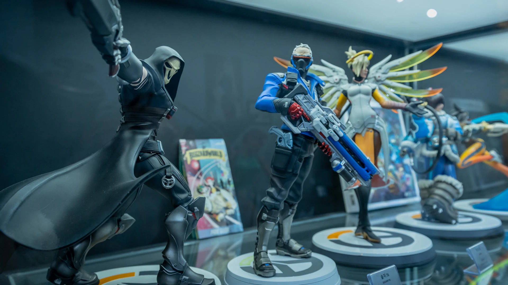 Three detailed figurines of characters from the game Overwatch displayed on stands, alongside a comic book in the background.