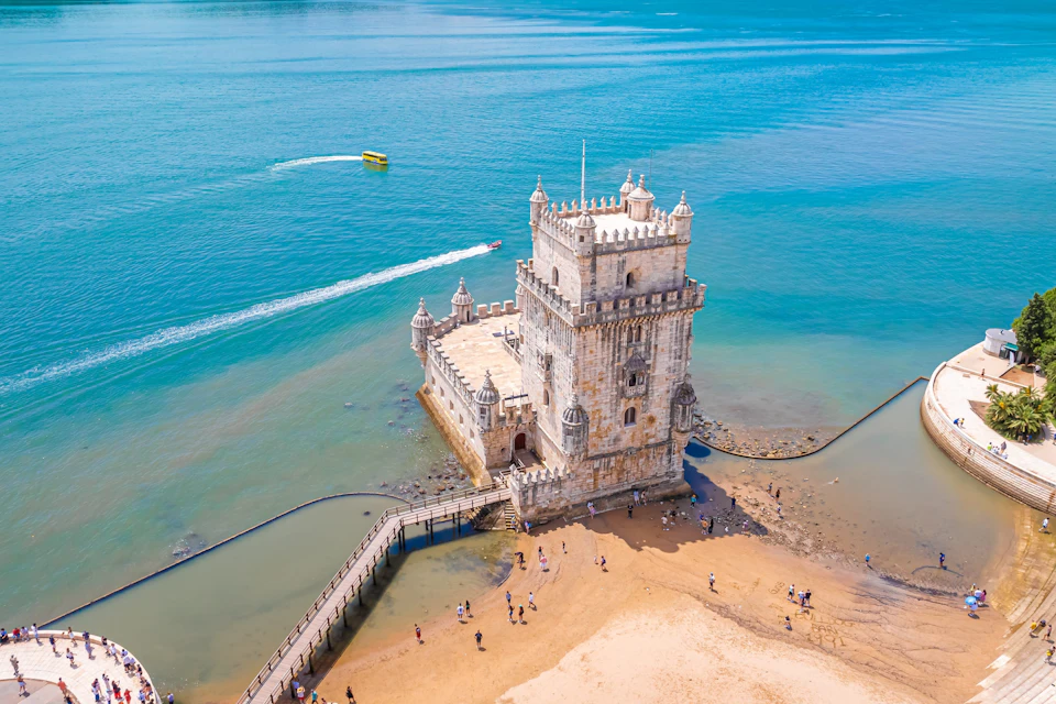 A historic tower on the coast surrounded by water, with people on the beach and a boat leaving a trail in the sea.