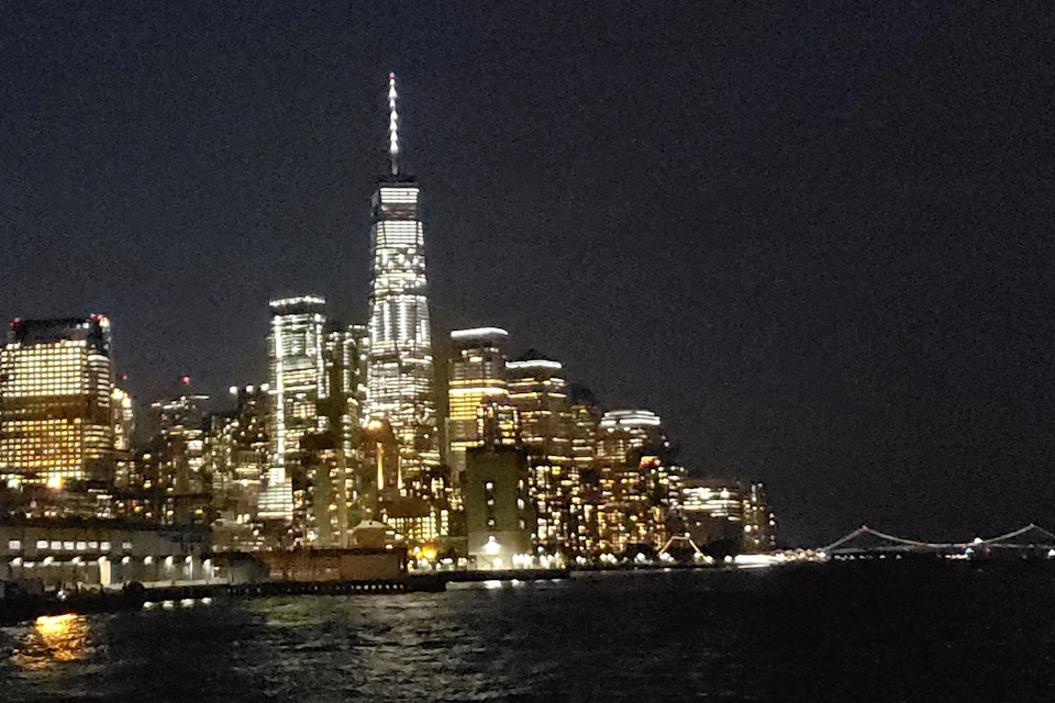 A city skyline at night with illuminated skyscrapers reflected on water, dominated by a tall, lit tower.