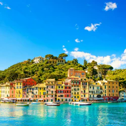 Färgglada byggnader kantar en strandpromenad med båtar förtöjda mot en kuliss av gröna kullar under en klarblå himmel.