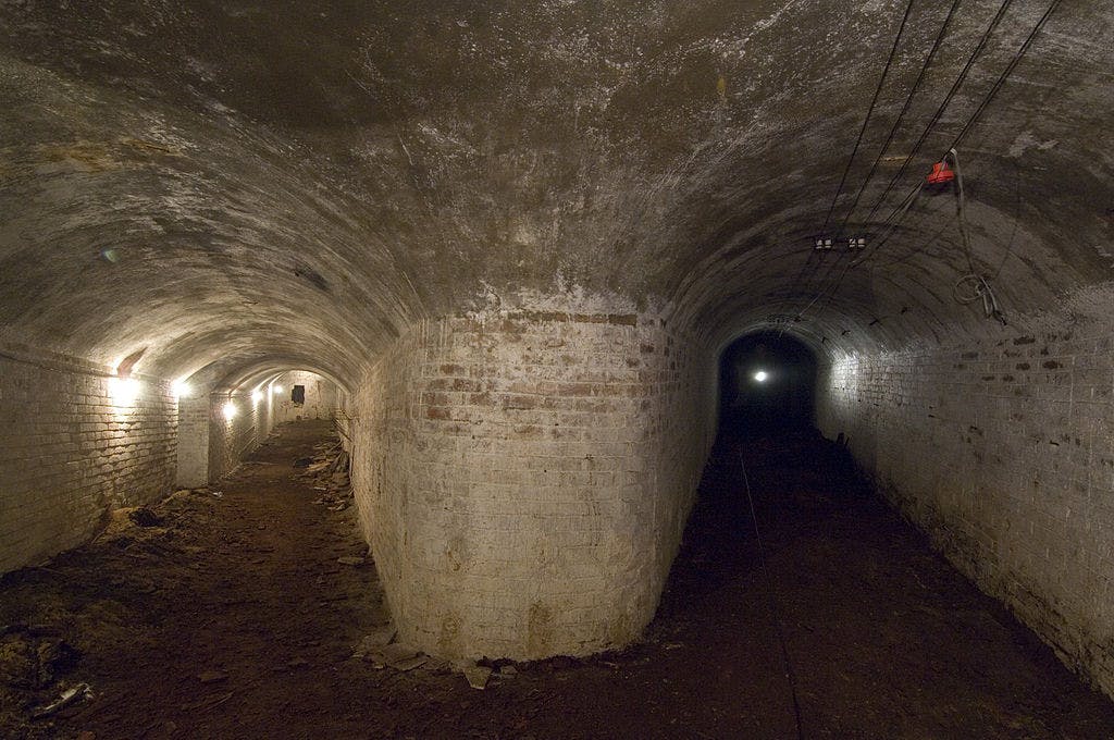 Underground brick tunnel with two diverging paths, lit by wall-mounted lights. Exposed wires run along the ceiling.
