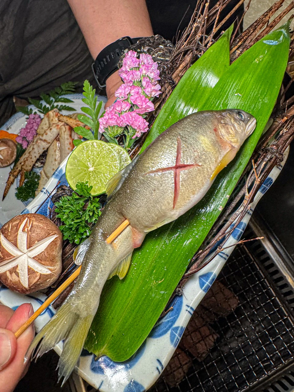 A prepared fish on a skewer placed on a green leaf, accompanied by mushrooms, lime, and flowers on a decorative platter.