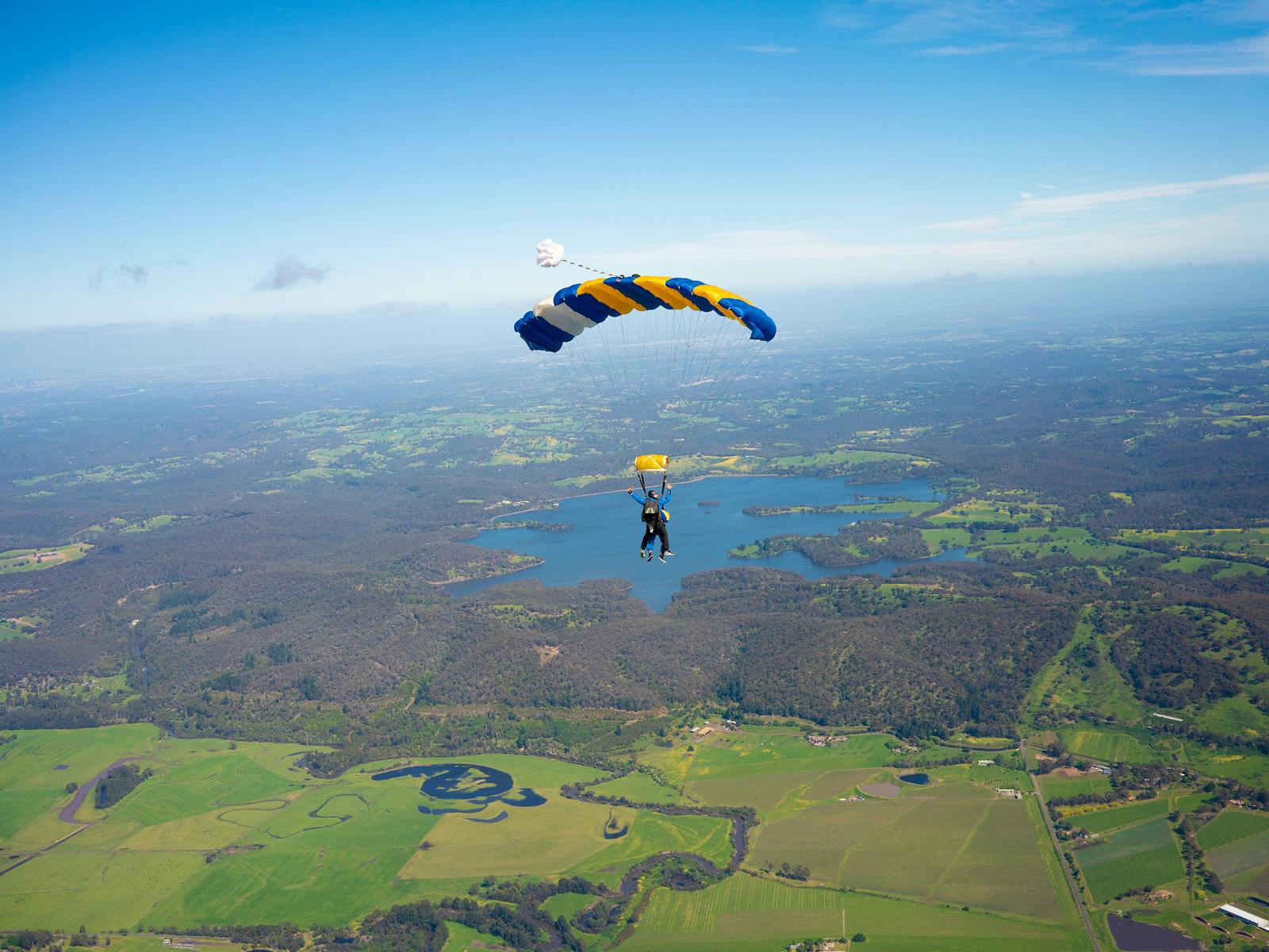 A person with a blue and yellow parachute descends over a vast landscape of fields and a lake on a clear day.