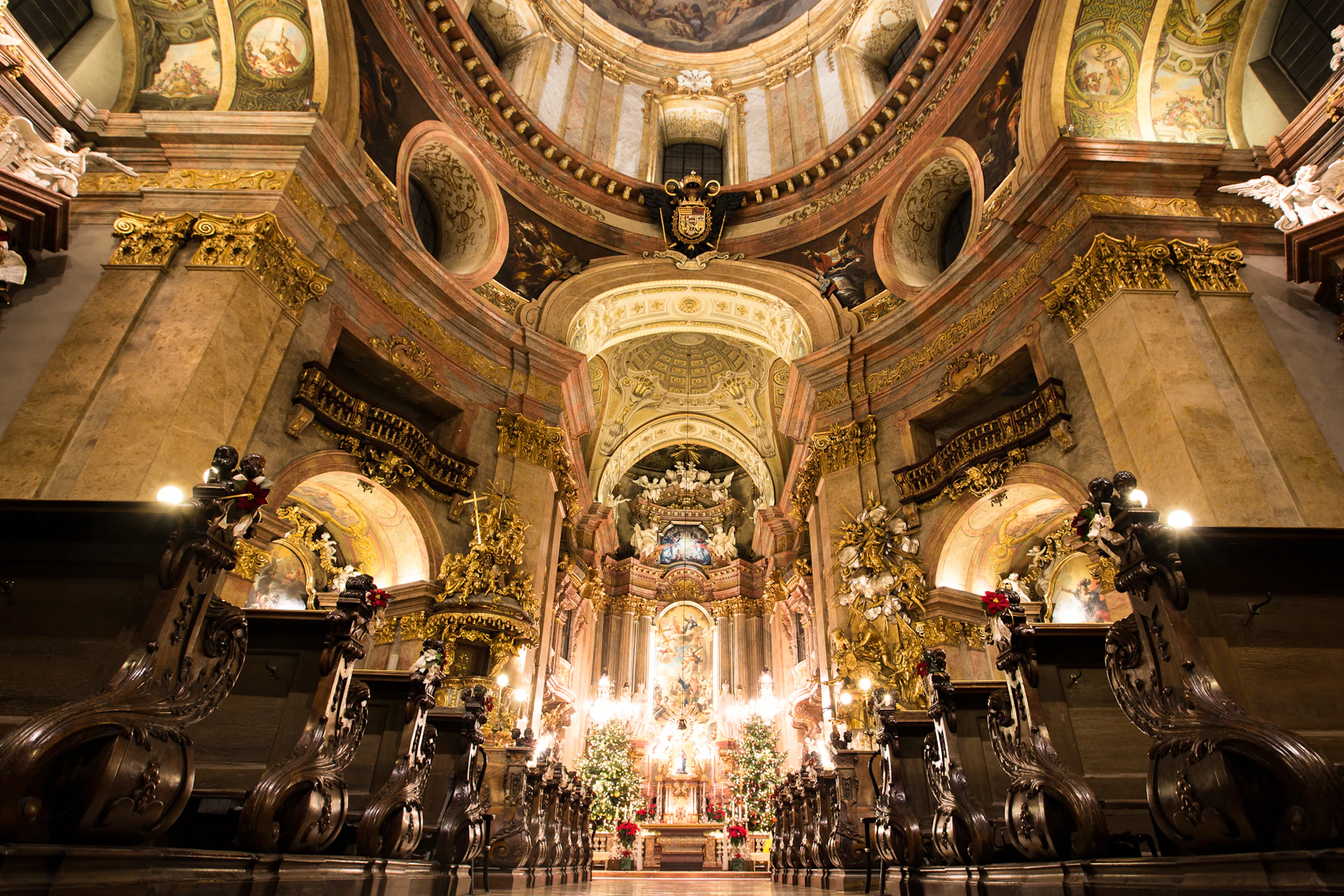 Ornate church interior with golden decorations, high ceilings, chandeliers, and wooden pews, focusing on a central altar with religious statues.