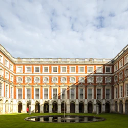 Symmetrical courtyard with a round central fountain, surrounded by a red-brick building with arched windows under a partly cloudy sky.