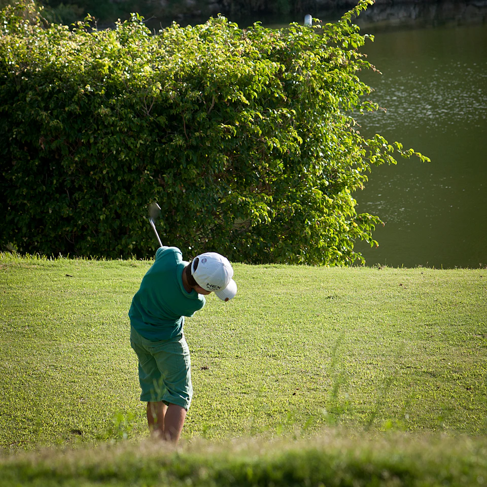 Person in a green outfit playing golf, swinging a club on a grassy course with a lake and bushes in the background.