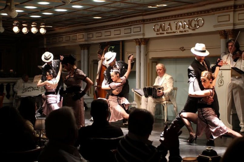 Couples dressed in formal attire perform a tango dance on stage while a band plays music at a venue named "Gala Tango."
